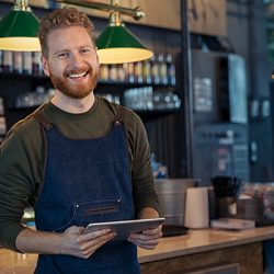 Successful small business owner using digital tablet and looking at camera. Happy smiling waiter wearing apron and holding digital tablet ready to take order. Portrait of young entrepreneur of coffee shop standing at counter with copy space.
